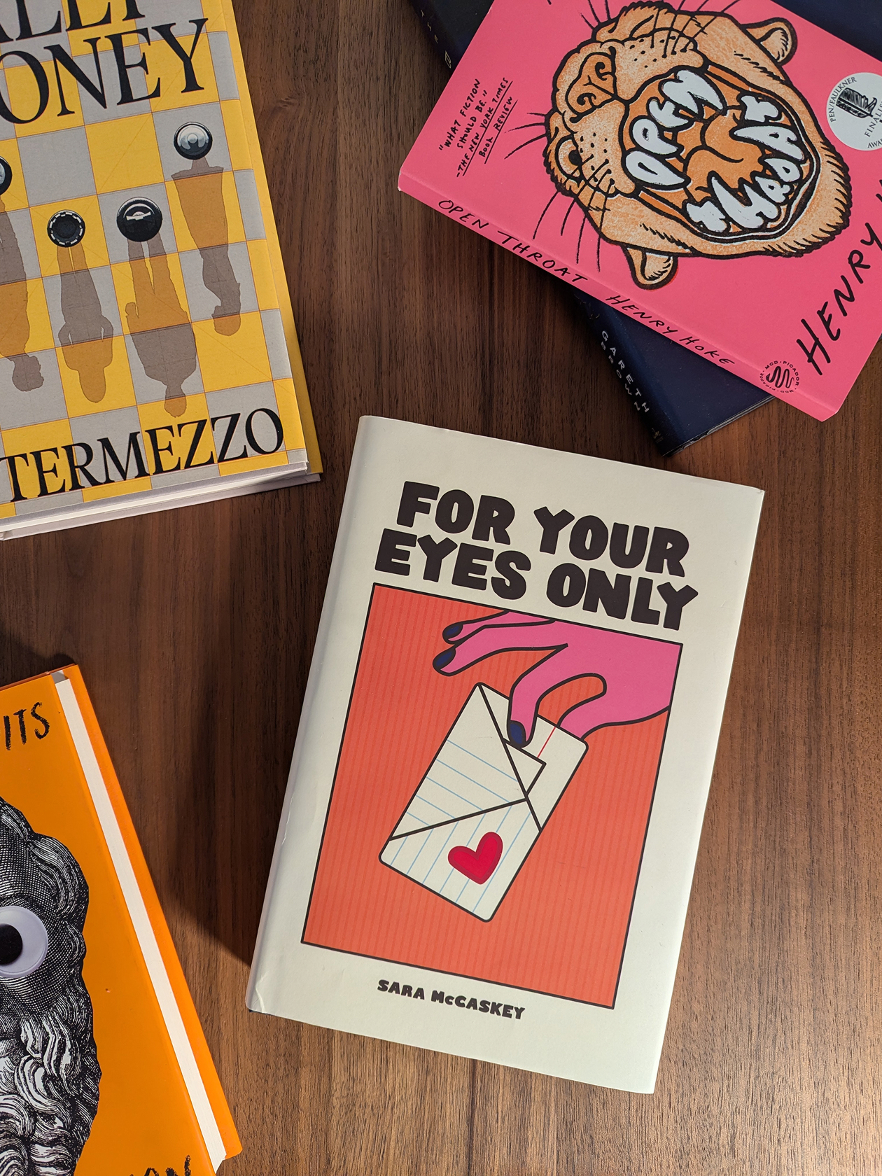 Sara’s debut novel For Your Eyes Only. A hardcover book showing an illustrated hand holding a folded paper note.
