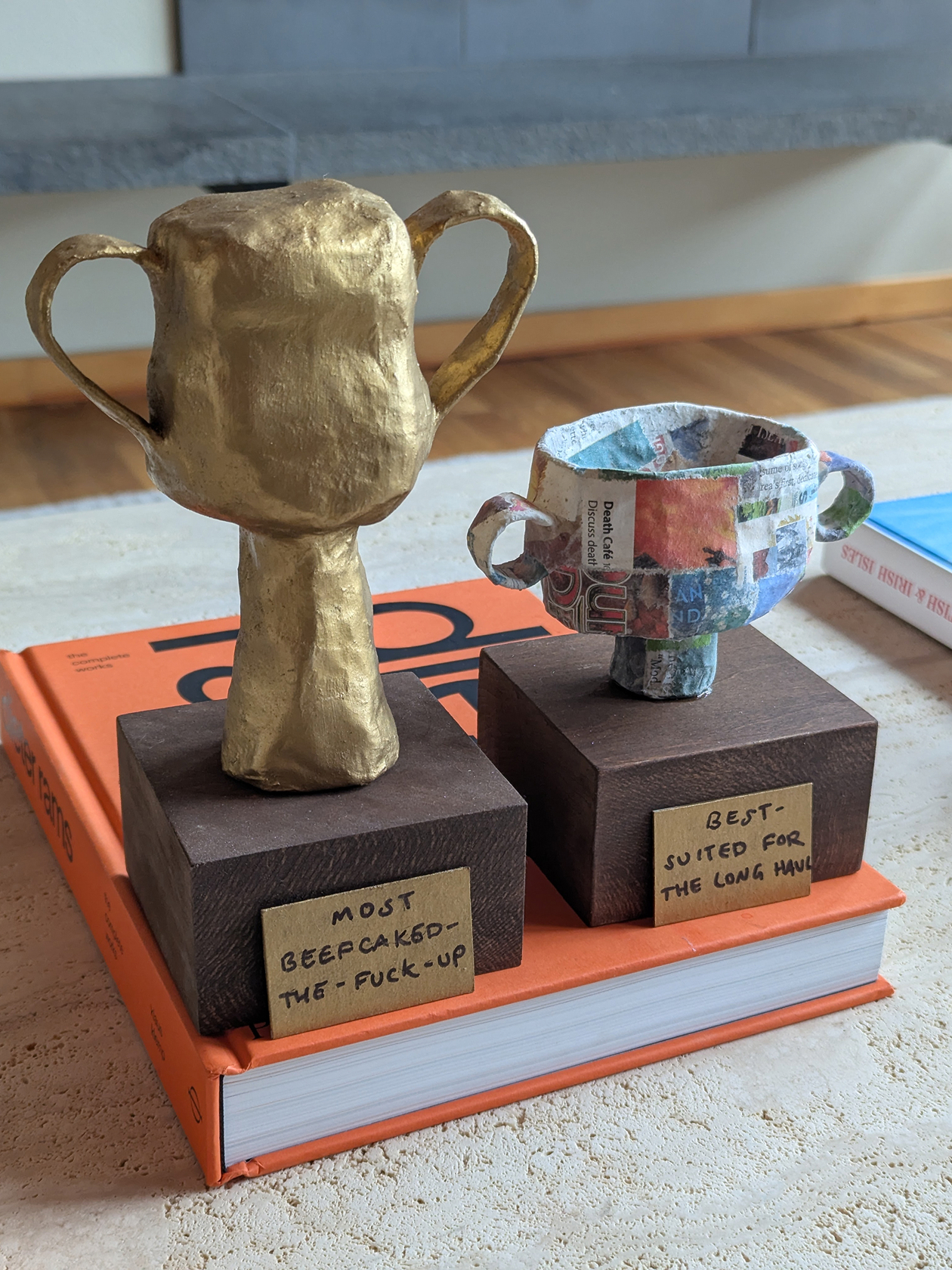 Two handmade paper mache trophies, one with a painted gold surface and the other with a patchwork newspaper collage on stained wood blocks with handwritten paper plaques.